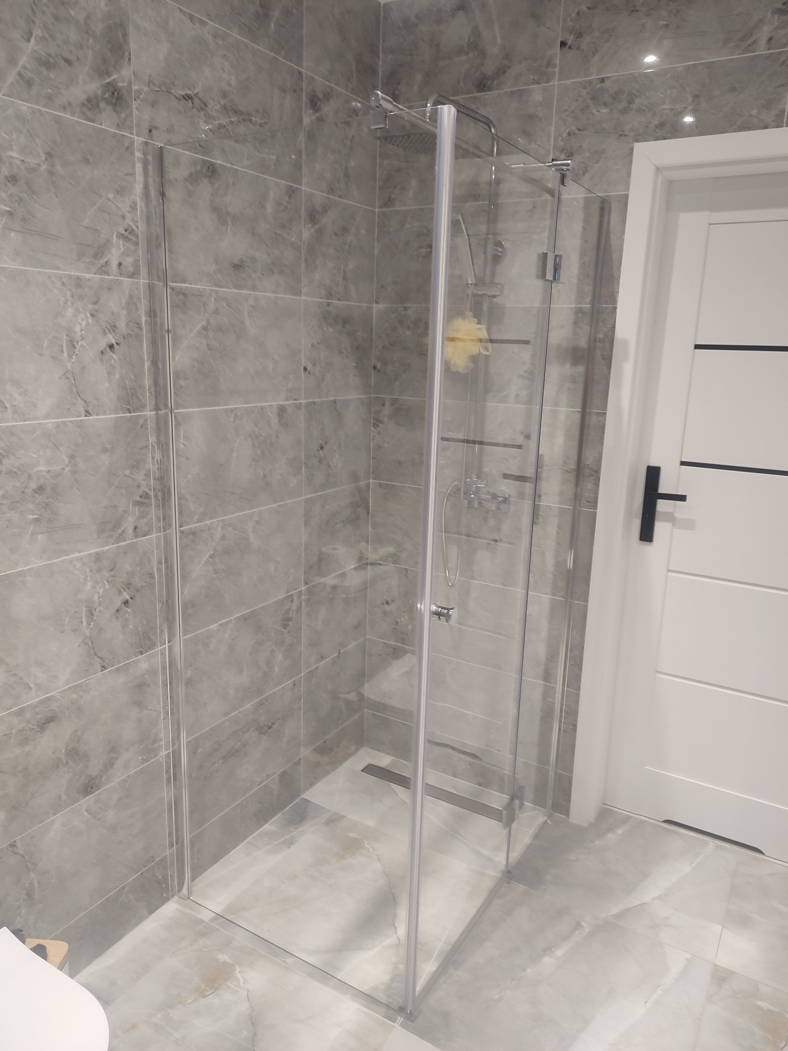 Szklana kabina prysznicowa z odpływem liniowym, wykończona płytkami imitującymi marmur. Białe drzwi z czarnym akcentem. Nowoczesna łazienka po remoncie.