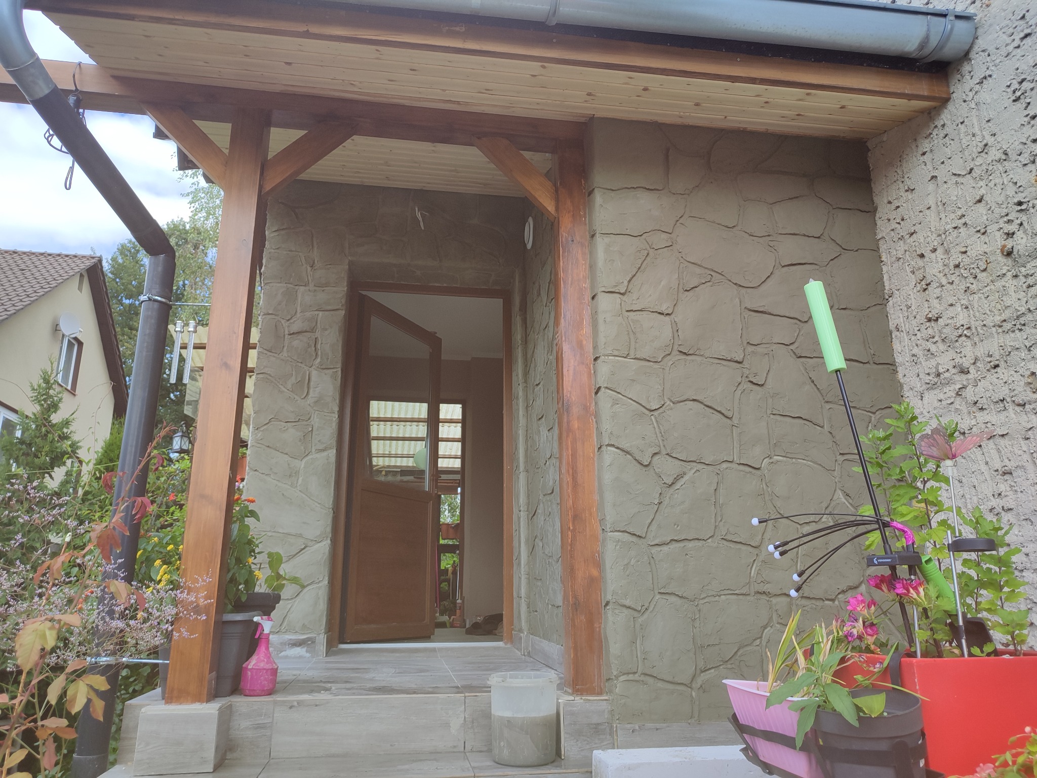 Dobudówka domu z drewnianą konstrukcją zadaszenia i elewacją imitującą kamień. Widoczne rośliny w donicach i częściowo otwarte drzwi.
