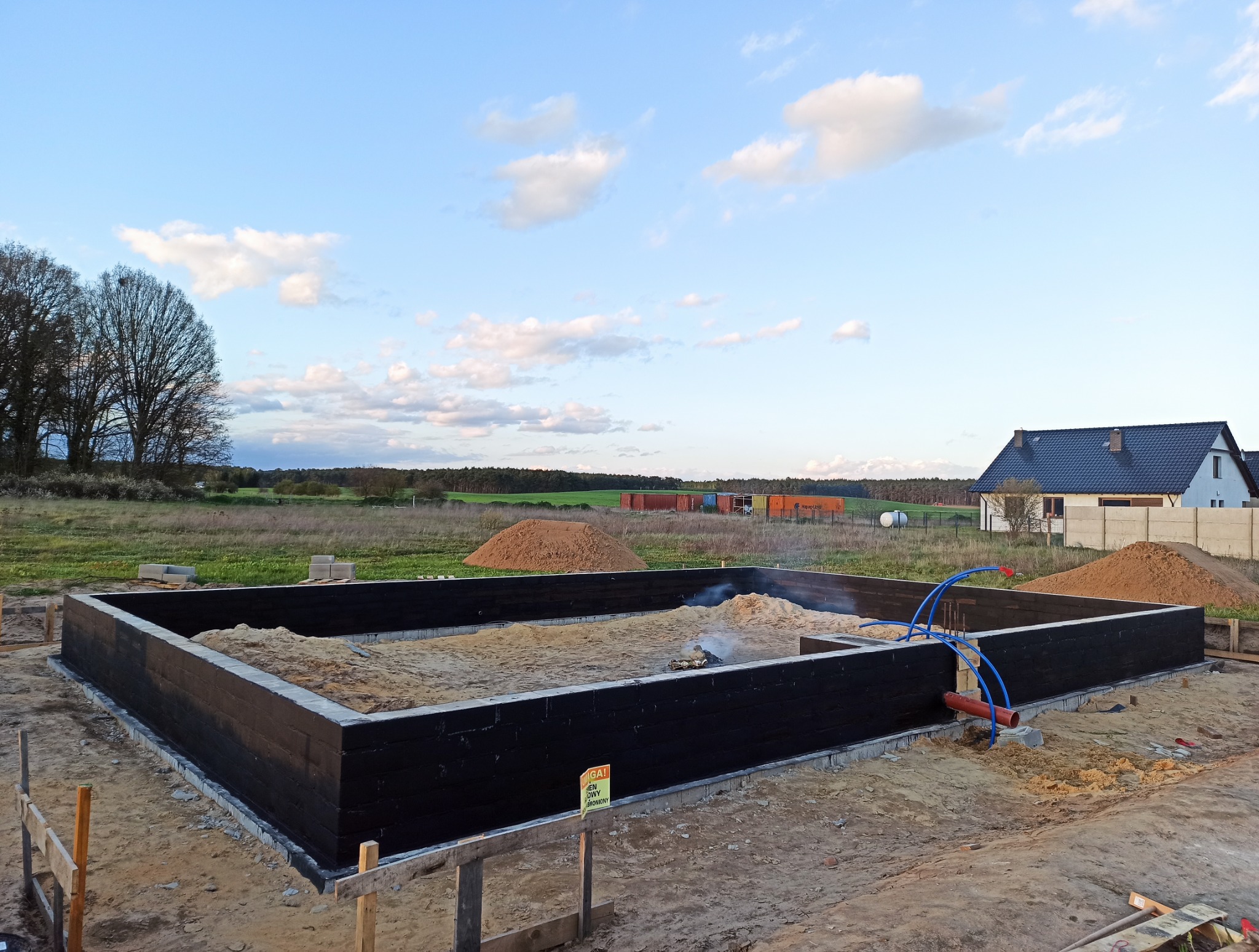 Zalane fundamenty domu jednorodzinnego, zabezpieczone czarną izolacją, z widocznymi instalacjami wewnątrz. W tle dom i krajobraz.