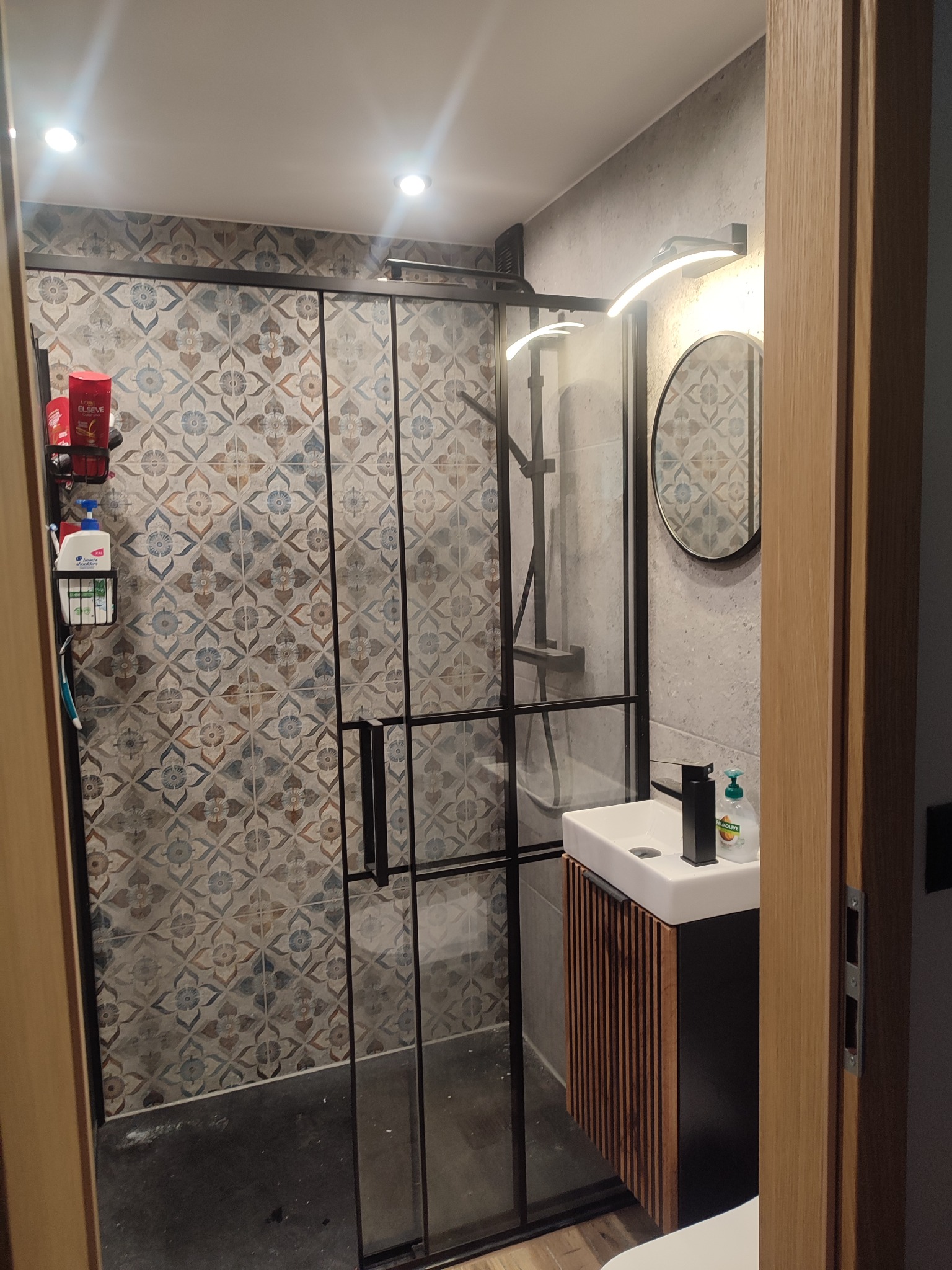 Nowoczesna łazienka z kabiną prysznicową typu walk-in, wzorzyste płytki, umywalka z szafką z lamelami i okrągłe lustro z oświetleniem LED.