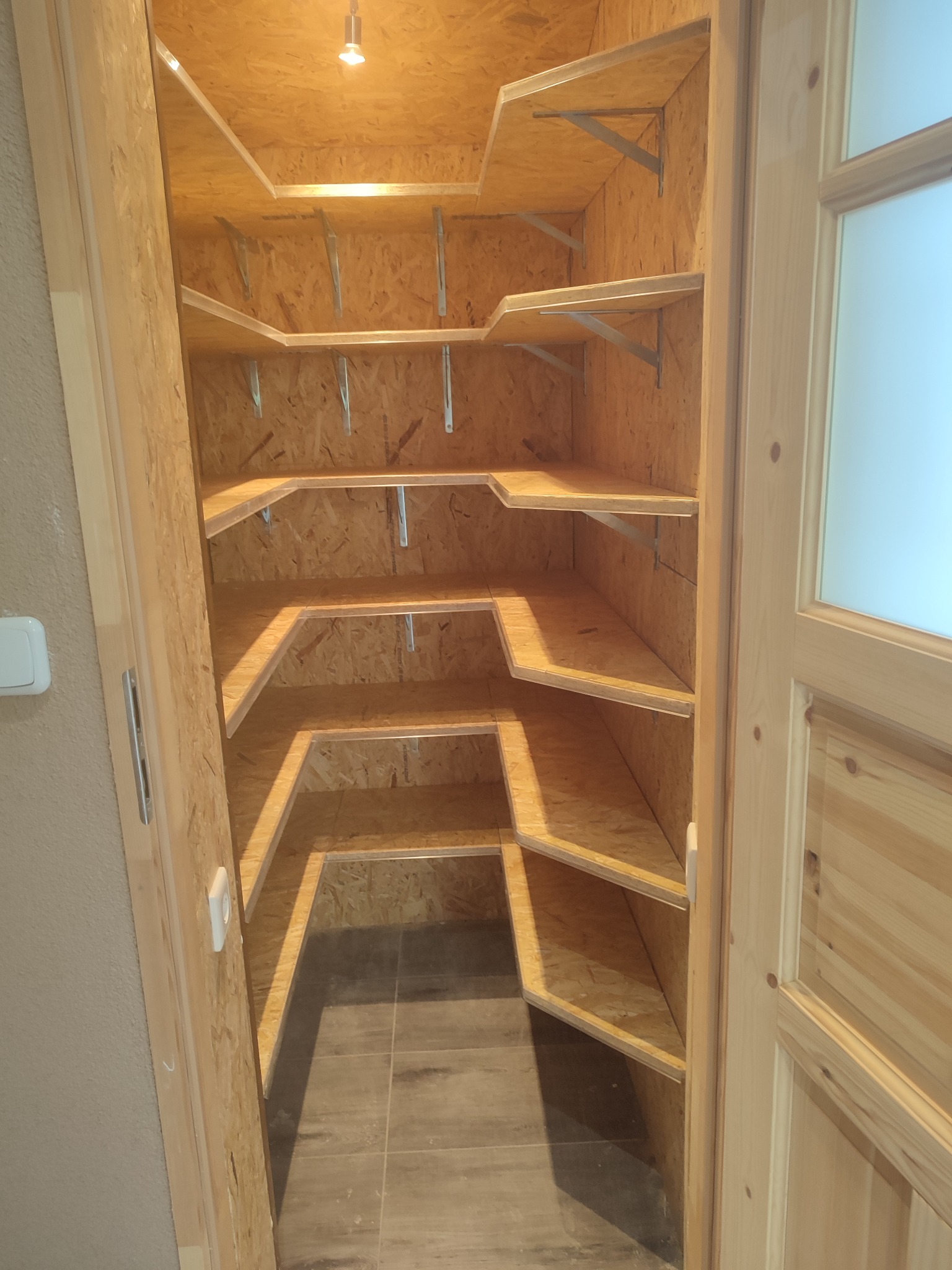 Wykonana z płyty OSB spiżarnia z półkami, oświetlona punktowym światłem. Widoczne metalowe wsporniki półek i wykończenie z surowego drewna. Podłoga wyłożona płytkami.