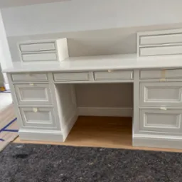 Białe biurko z szufladami w klasycznym stylu, z naklejoną taśmą malarską na frontach, umieszczone na drewnianej podłodze zabezpieczonej kartonem i folią.