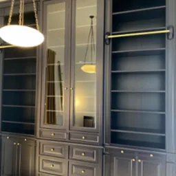 Elegancka, ciemna zabudowa meblowa z przeszklonymi witrynami i złotymi akcentami, oświetlona lampą wiszącą. Widoczne szuflady i półki.