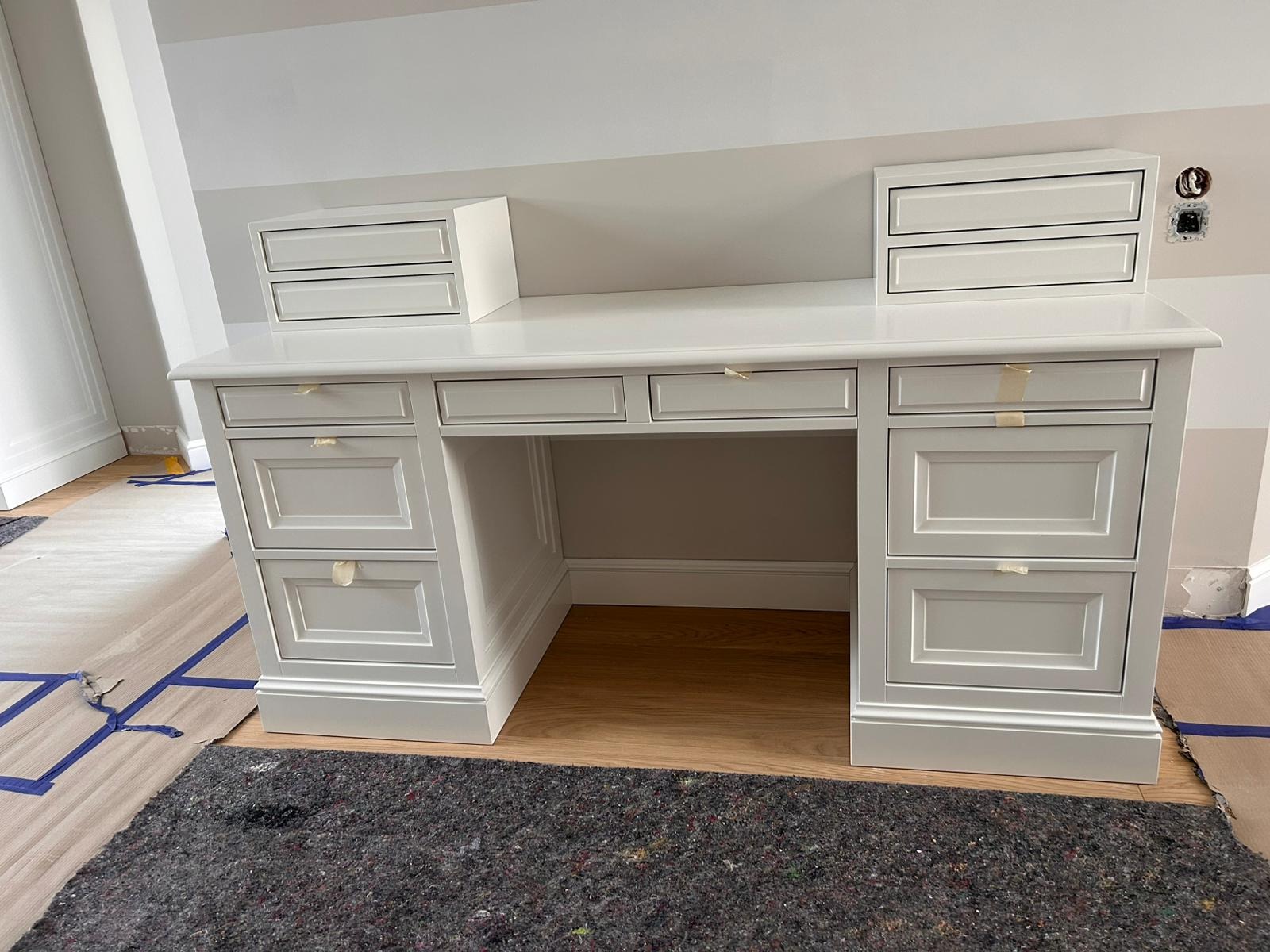 Białe biurko z szufladami w klasycznym stylu, z naklejoną taśmą malarską na frontach, umieszczone na drewnianej podłodze zabezpieczonej kartonem i folią.