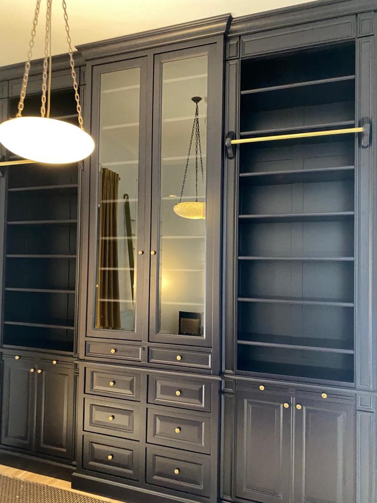 Elegancka, ciemna zabudowa meblowa z przeszklonymi witrynami i złotymi akcentami, oświetlona lampą wiszącą. Widoczne szuflady i półki.
