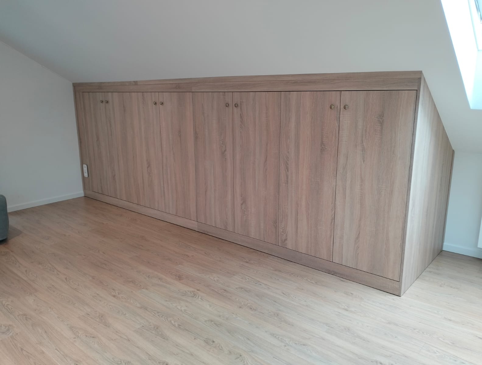 Jasna, drewnopodobna szafa wnękowa na poddaszu, z widocznymi drzwiami na zawiasach i podłogą z paneli. Dopasowanie do skosu dachu.