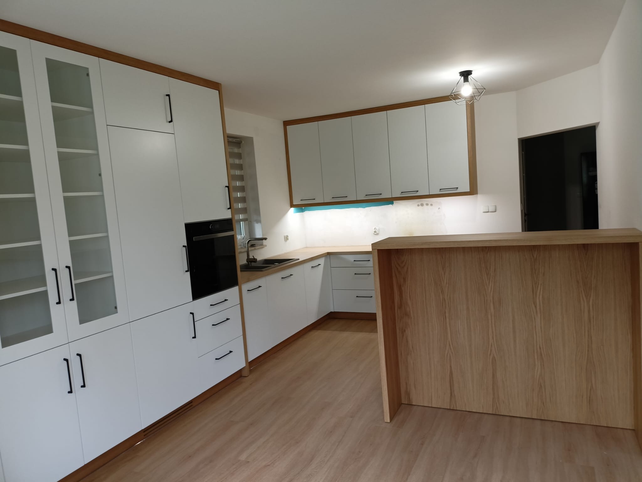 Nowoczesna kuchnia z białymi frontami, drewnianymi akcentami i czarnymi uchwytami. Widoczny barek i podświetlenie LED pod szafkami wiszącymi. Podłoga z paneli.