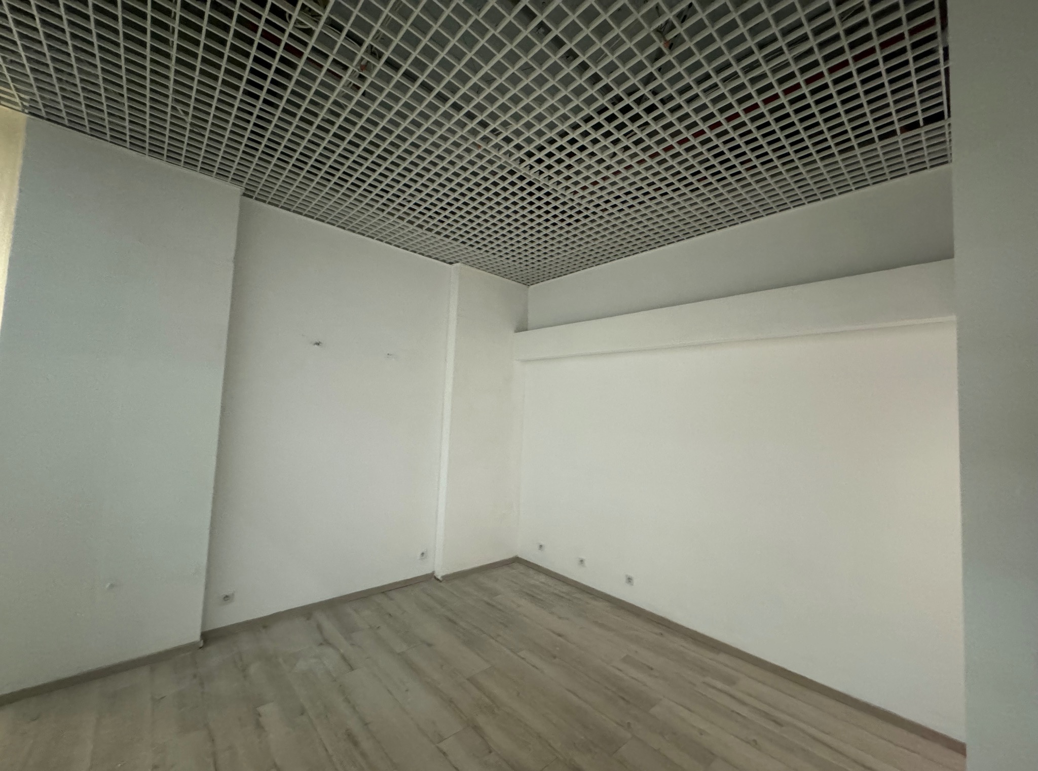 Puste, jasne pomieszczenie biurowe z podwieszanym sufitem rastrowym i panelami podłogowymi imitującymi drewno. Świeżo pomalowane ściany z widocznymi gniazdkami elektrycznymi.