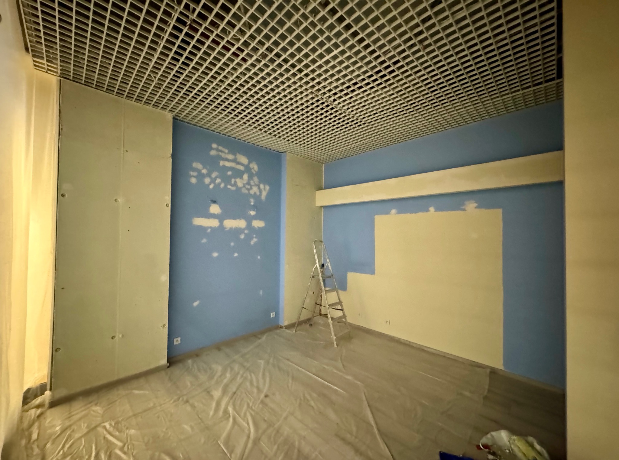 Wnętrze pokoju w trakcie malowania: niebieskie ściany z fragmentami szpachli, częściowo pomalowane na kremowo. Na podłodze folia ochronna, w rogu drabina.