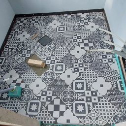 Usługi Ogólnobudowlane Andrzej - Układanie wzorzystych, czarno-białych płytek podłogowych w łazience. Widoczne narzędzia i fragment instalacji. Prace w toku, etap wykończeniowy.