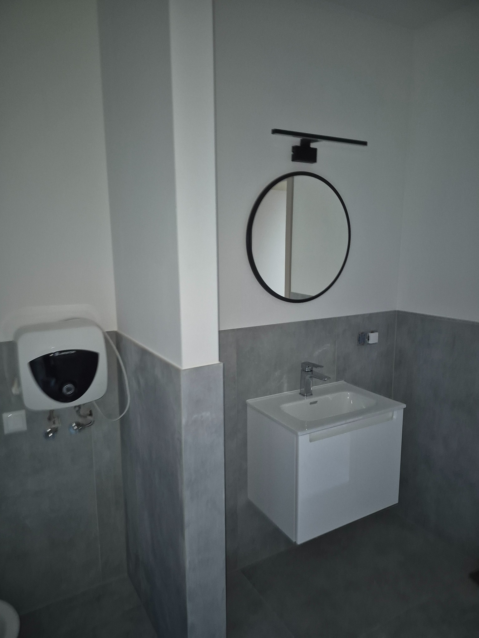 Nowoczesna łazienka z okrągłym lustrem, białą szafką umywalkową i szarymi płytkami do połowy ściany. Na ścianie bojler elektryczny. Minimalistyczny design.