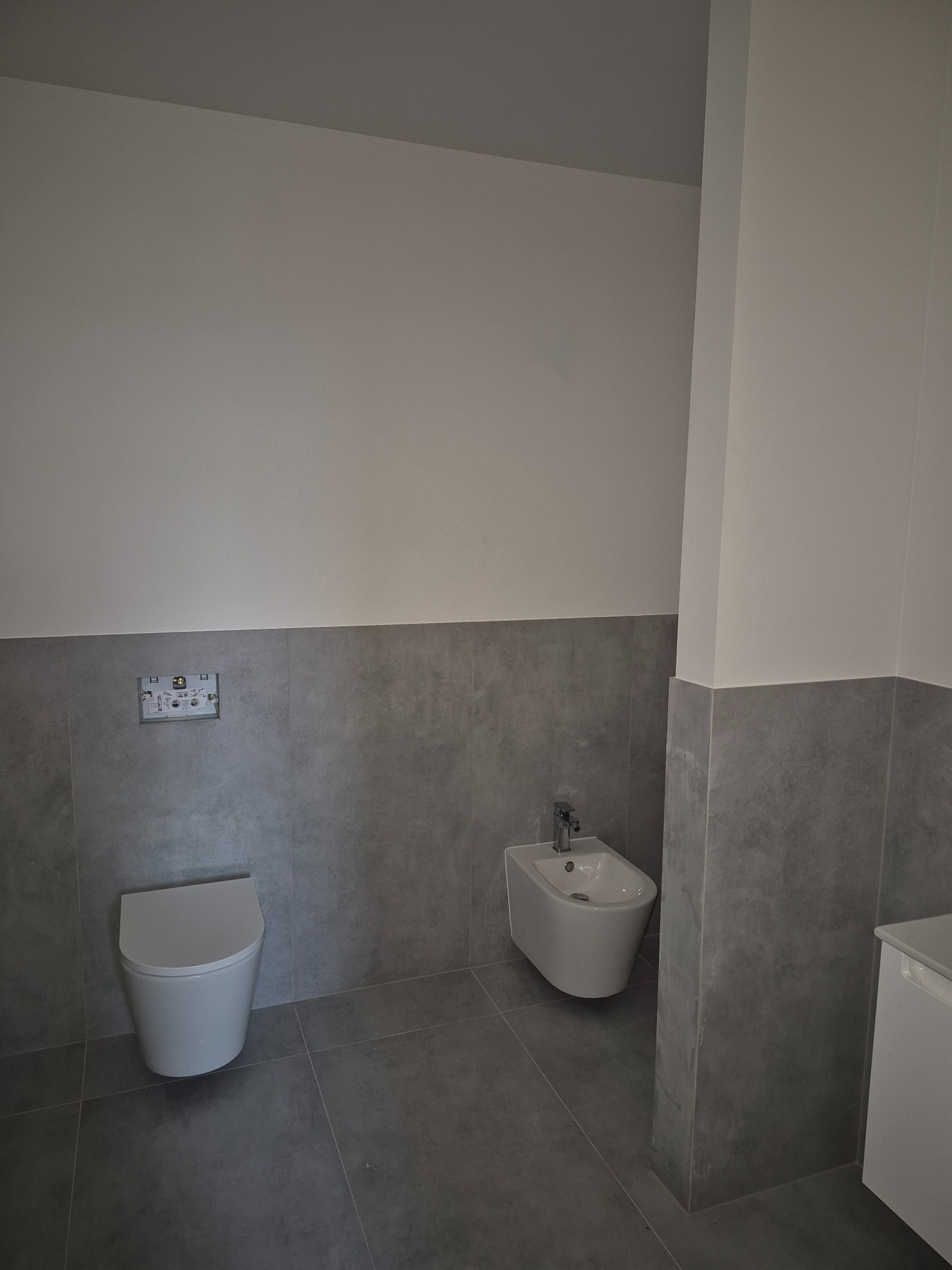 Nowoczesna łazienka z szarymi płytkami, wiszącą toaletą i bidetem. Minimalistyczny design, stonowana kolorystyka, widoczne elementy instalacji podtynkowej.