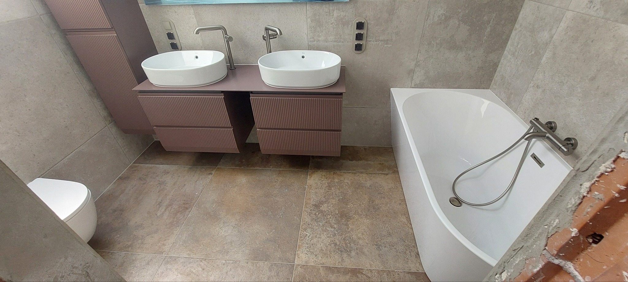 Nowoczesna łazienka z podwójną umywalką na szafce w odcieniach różu, białą wanną i beżowymi płytkami. Widoczny fragment toalety. Minimalistyczny design.