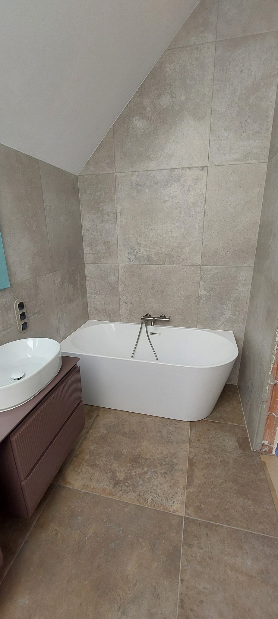 Nowoczesna łazienka z wanną, umywalką na szafce i beżowymi płytkami na ścianach i podłodze. Ukośny sufit dodaje unikalnego charakteru wnętrzu.