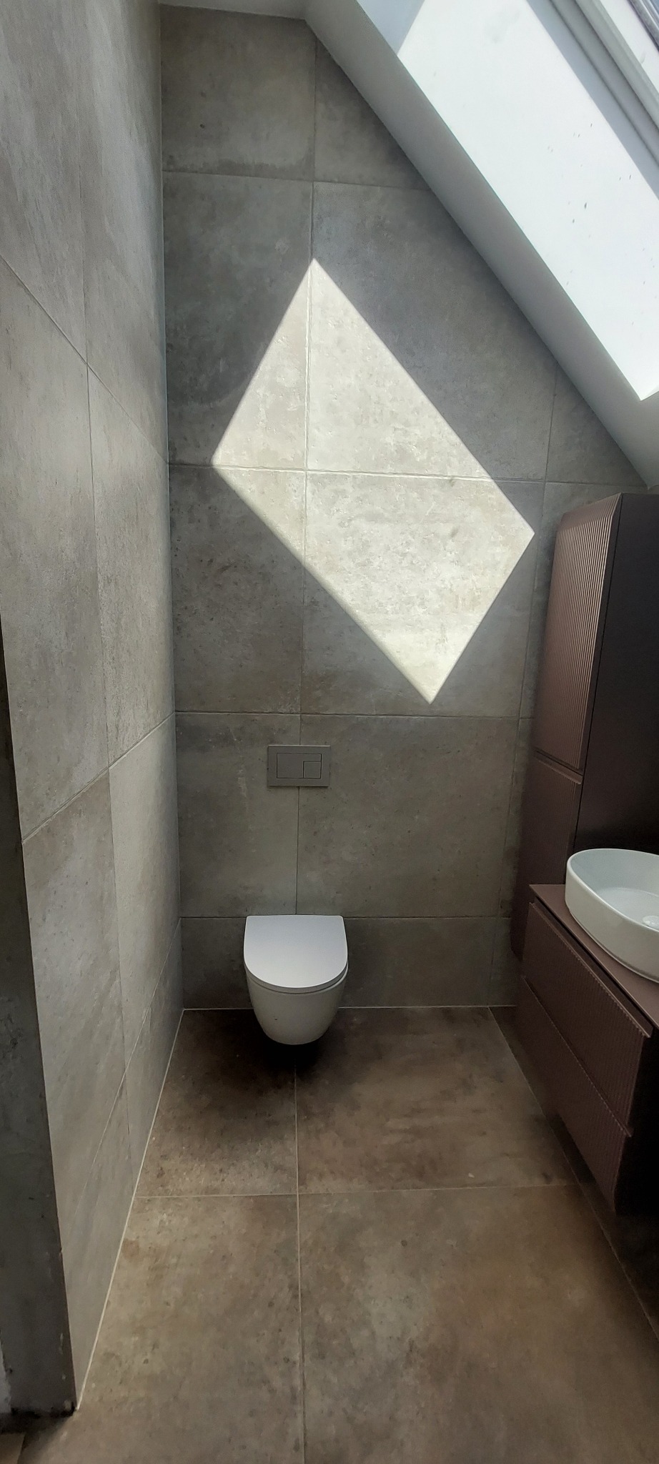 Wykończona łazienka na poddaszu z szarymi płytkami, wiszącą toaletą i szafką z umywalką. Uwagę przyciąga mocne światło słoneczne padające przez okno dachowe.