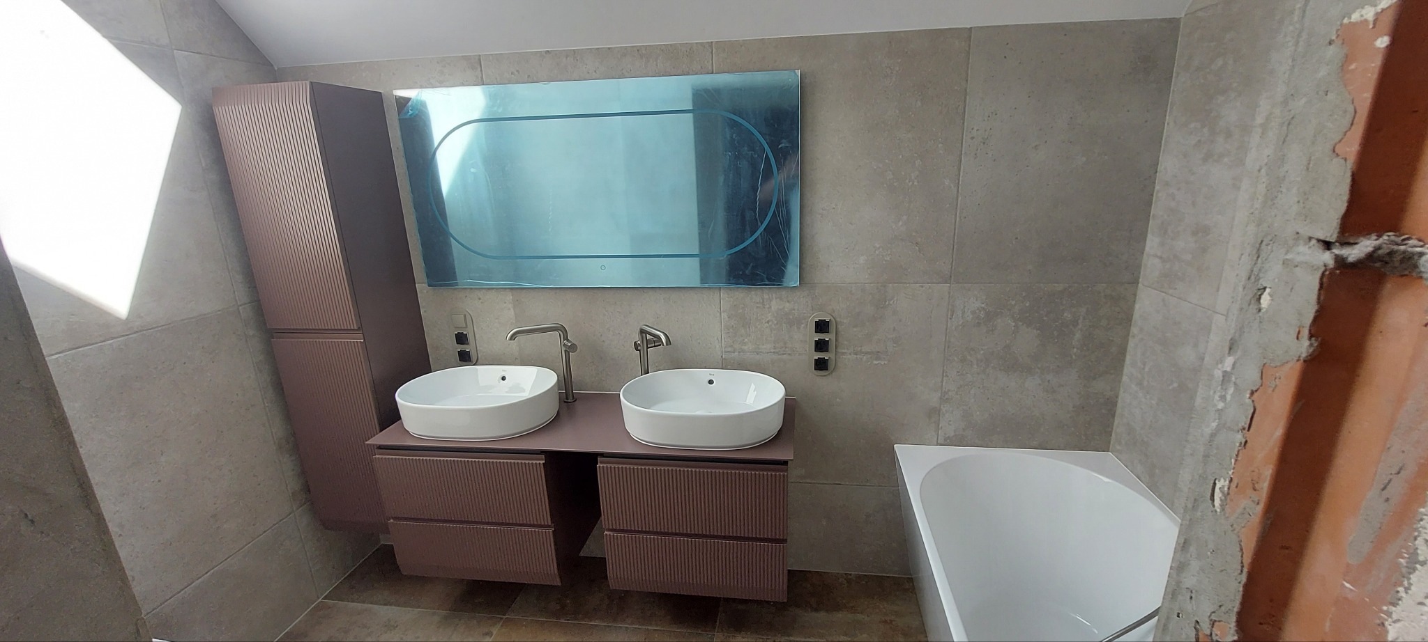 Nowoczesna łazienka z dwoma umywalkami, szafką w kolorze pudrowego różu i dużym lustrem z folią ochronną. Widoczna wanna i fragment ściany w trakcie wykończenia.