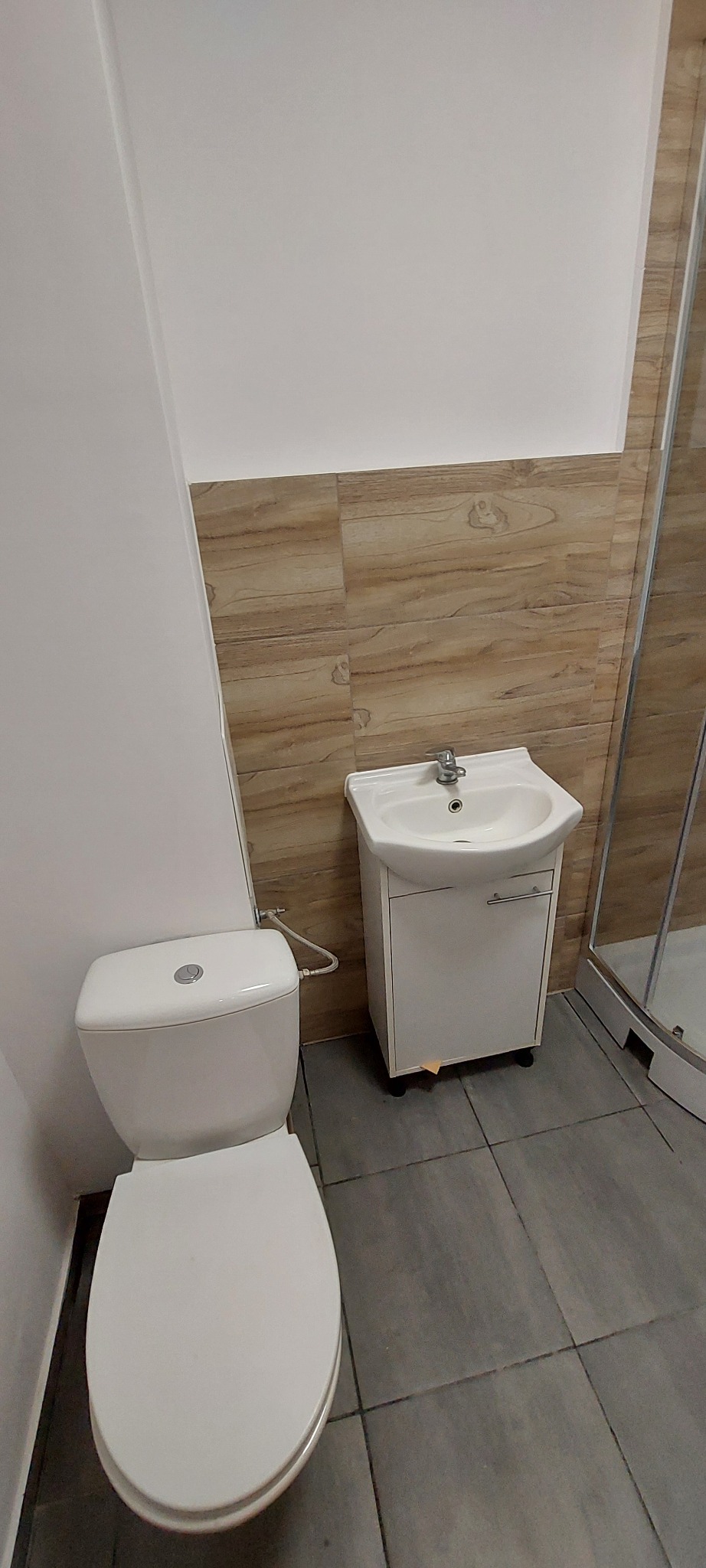 Wykończona łazienka: biała toaleta i umywalka z szafką na tle płytek imitujących drewno i szarej podłogi. Widoczny fragment kabiny prysznicowej.