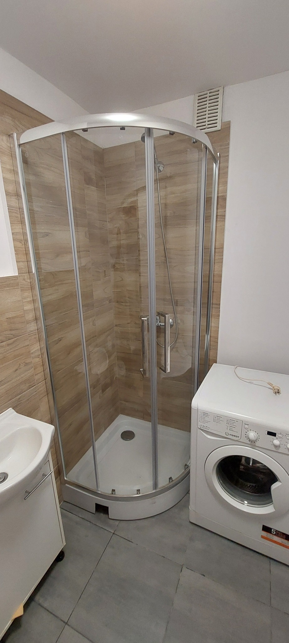 Nowoczesna kabina prysznicowa z drewnopodobnymi płytkami, umywalką i pralką w jasnej łazience. Szare płytki podłogowe, minimalistyczny design.