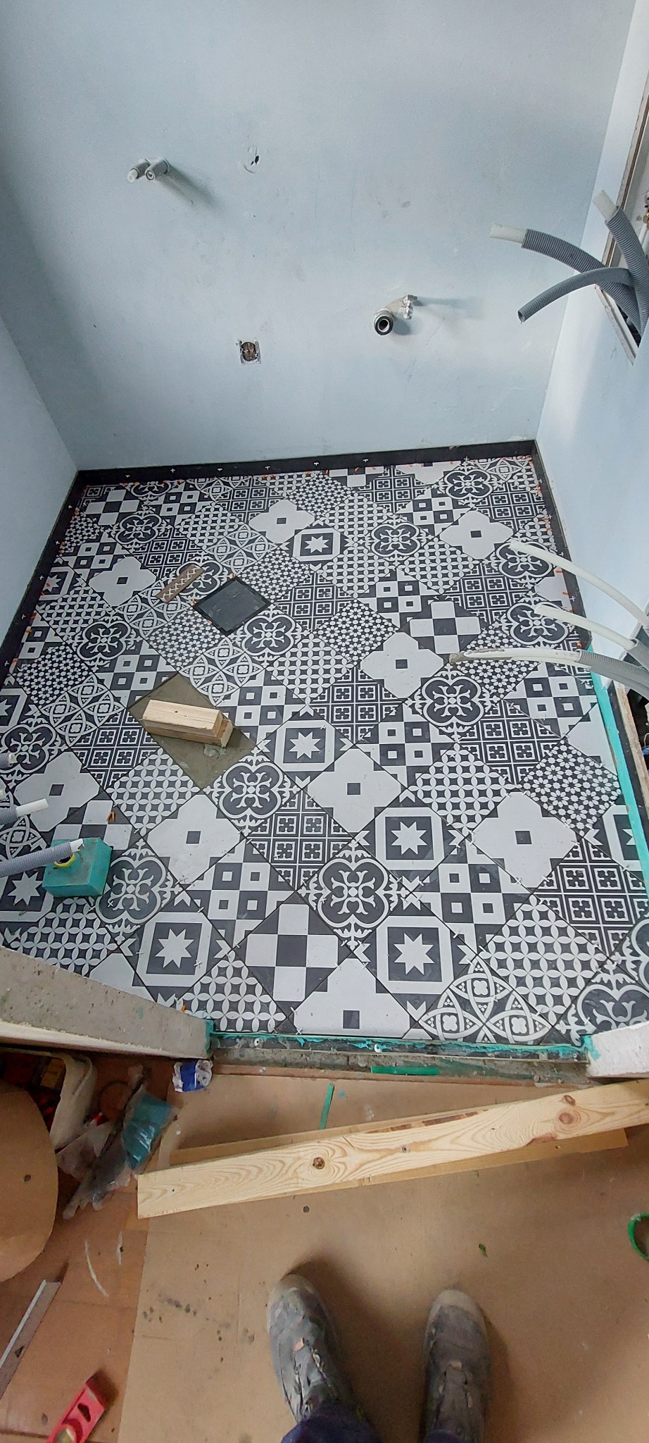 Układanie wzorzystych, czarno-białych płytek podłogowych w łazience. Widoczne narzędzia i fragment instalacji. Prace w toku, etap wykończeniowy.