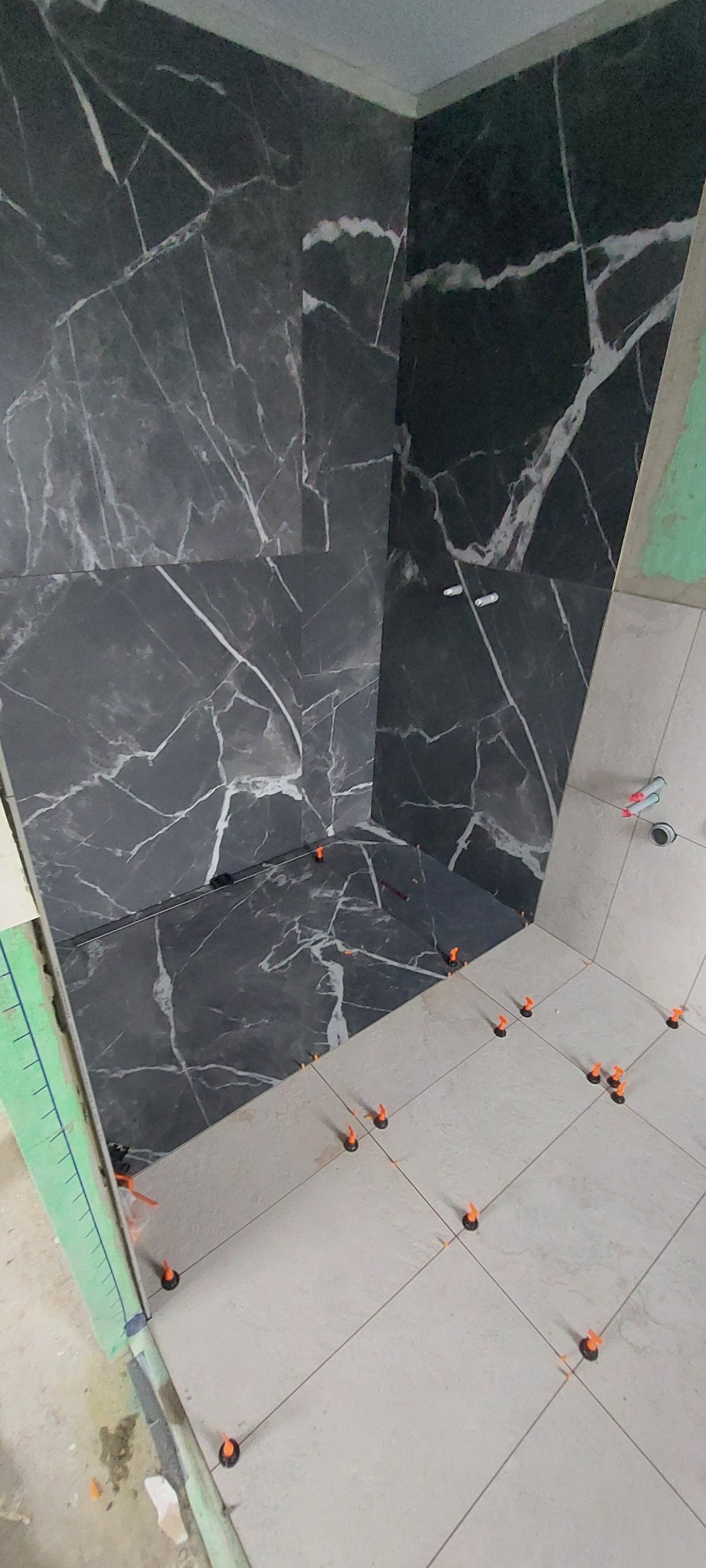 Układanie płytek imitujących marmur w kabinie prysznicowej. Widoczne pomarańczowe klipsy poziomujące. Częściowo wykończona podłoga z jasnych płytek.