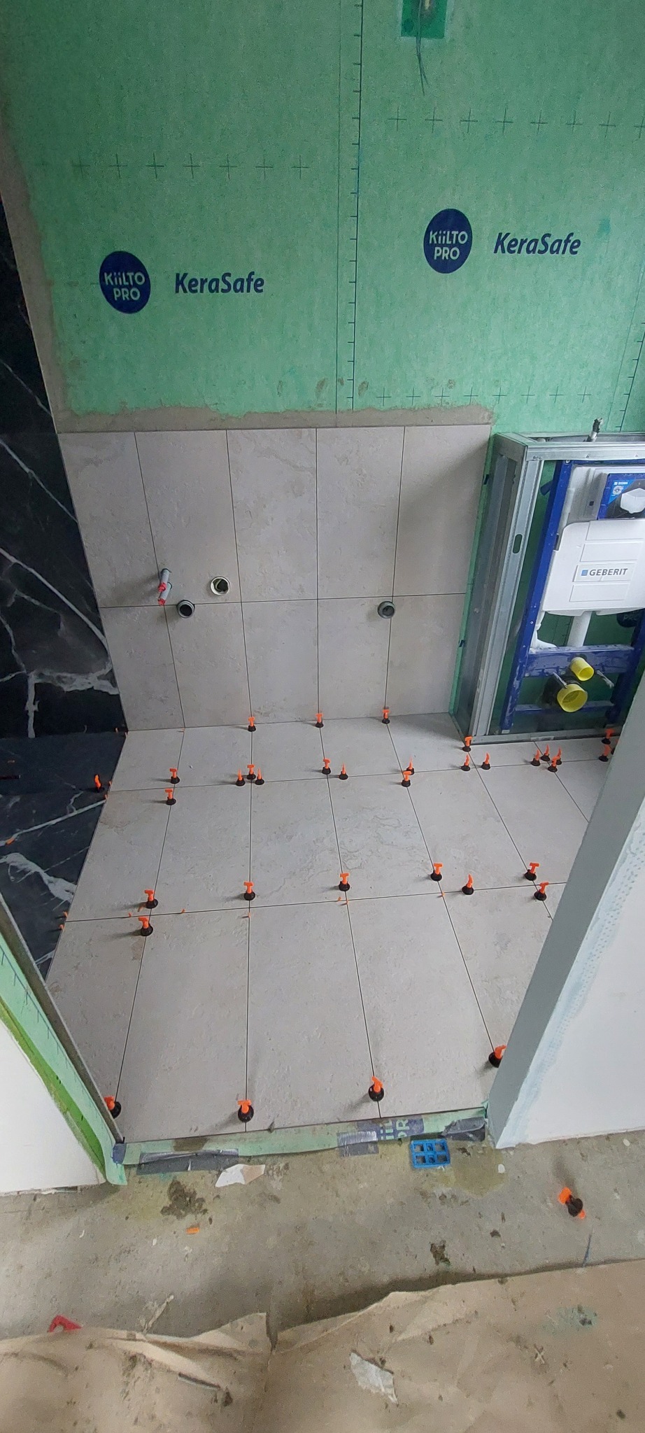 Układanie jasnych płytek w łazience z użyciem poziomujących klipsów. Widoczne płyty KeraSafe na ścianach i stelaż podtynkowy Geberit. Częściowo wykończona podłoga.