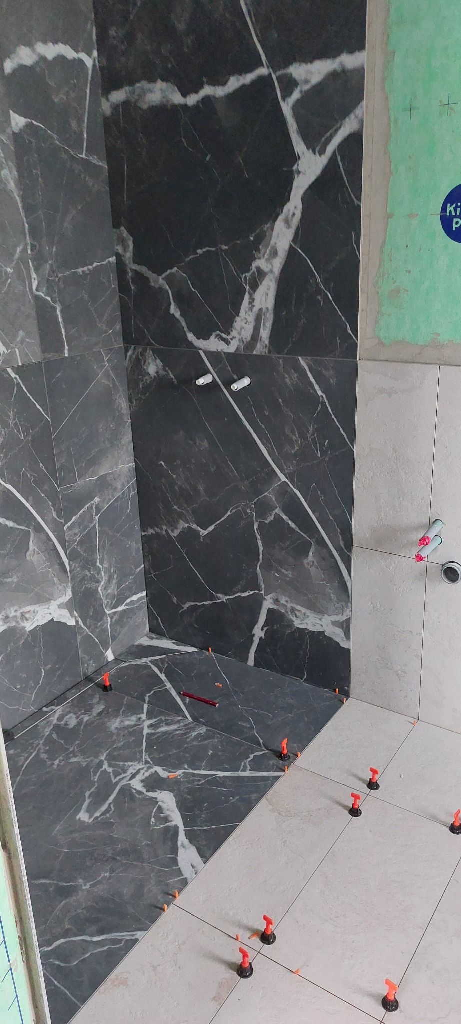 Częściowo wykończona łazienka z szarymi płytkami imitującymi marmur na ścianach i jasnymi płytkami na podłodze. Widoczne systemy poziomowania płytek.