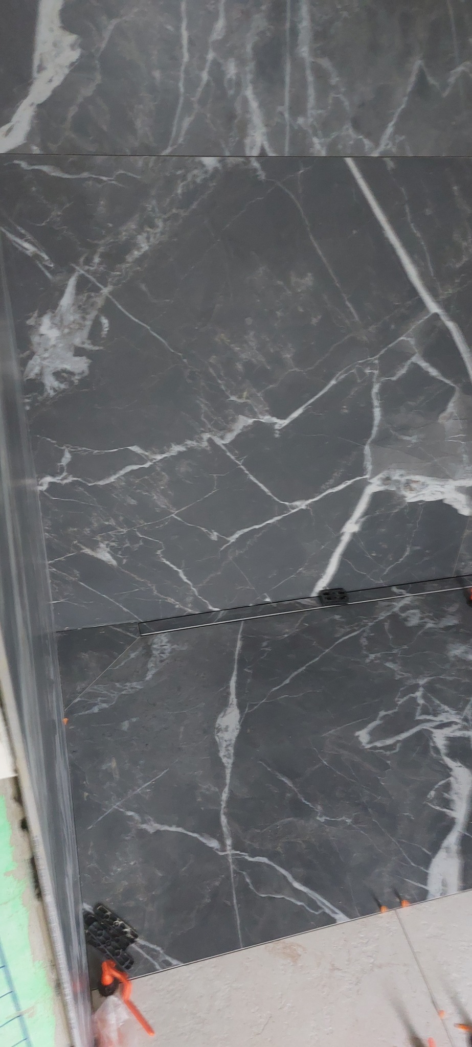 Ułożone, szare płytki imitujące marmur z białymi żyłami, widoczne czarne kliny dystansowe i odpływ liniowy. Fragment wykończonej podłogi w łazience.