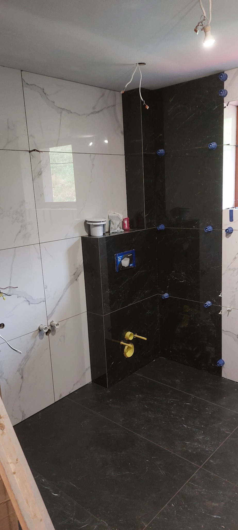 Częściowo wykończona łazienka z czarno-białymi płytkami imitującymi marmur, widoczne elementy instalacji i akcesoria glazurnicze.