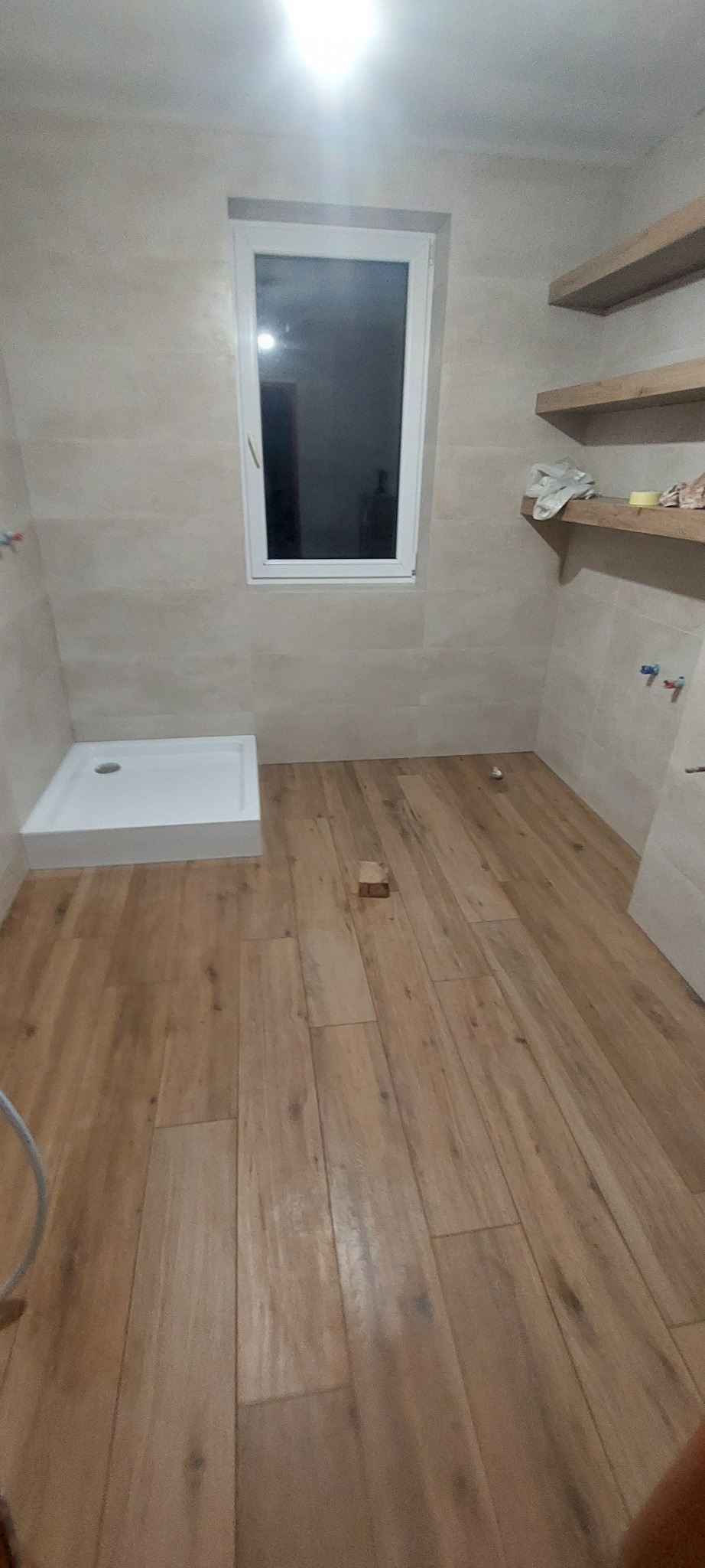 Wykończona łazienka z drewnopodobną podłogą, beżowymi płytkami ściennymi, białym brodzikiem i drewnianymi półkami. Widoczne przyłącza wodne.