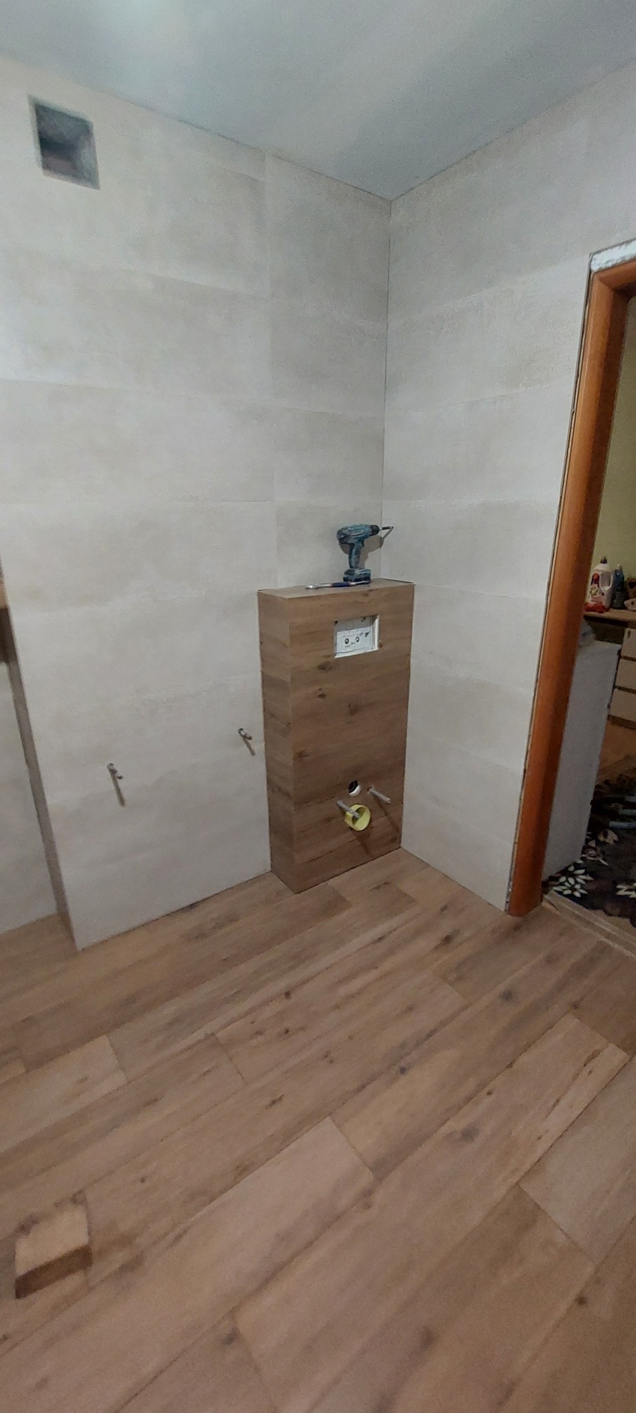 Wnętrze łazienki w trakcie wykańczania: zabudowa WC z drewnopodobną okleiną, płytki na ścianach, drewniana podłoga, wiertarka na obudowie stelaża.
