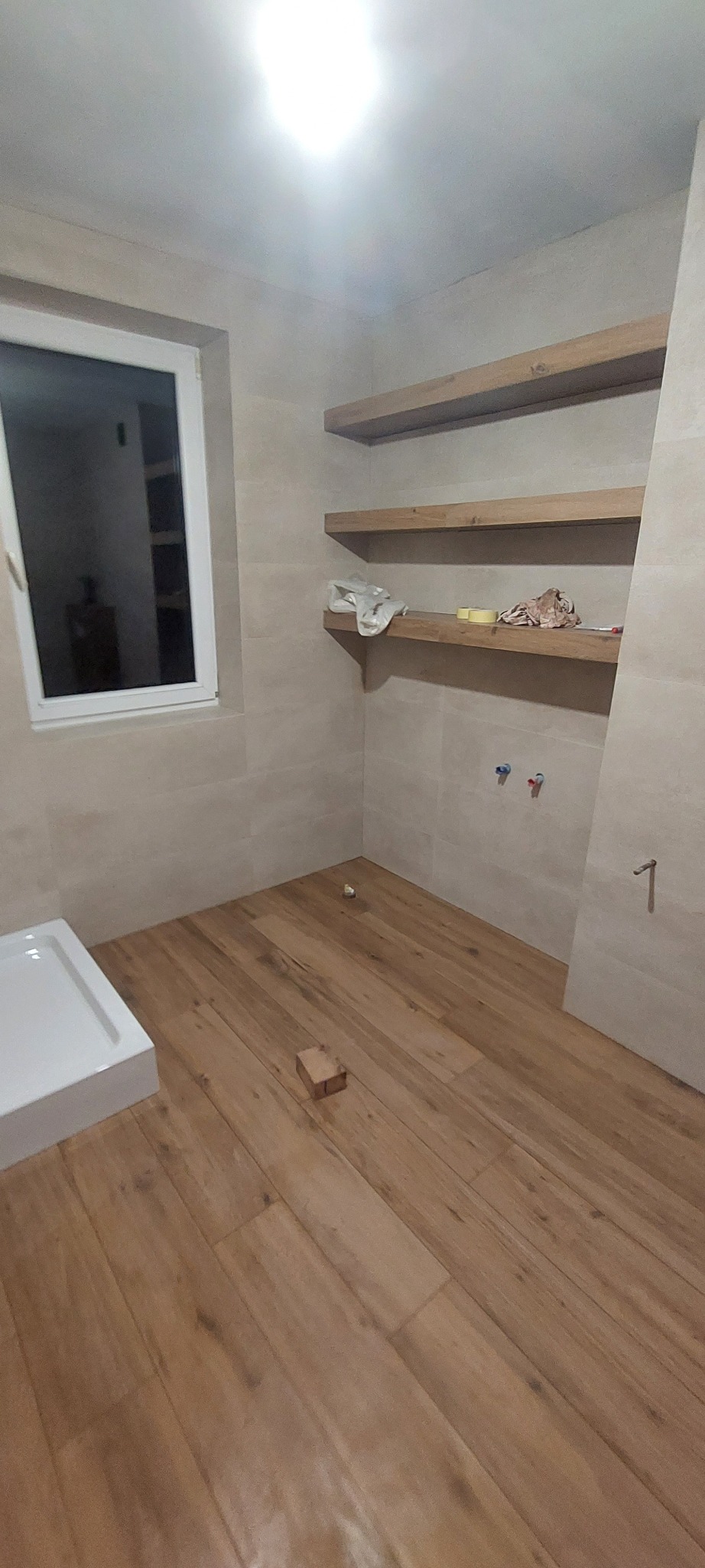 Jasna łazienka w trakcie wykańczania: drewnopodobne płytki na podłodze, beżowe płytki na ścianach, drewniane półki, biała rama okienna i fragment brodzika.