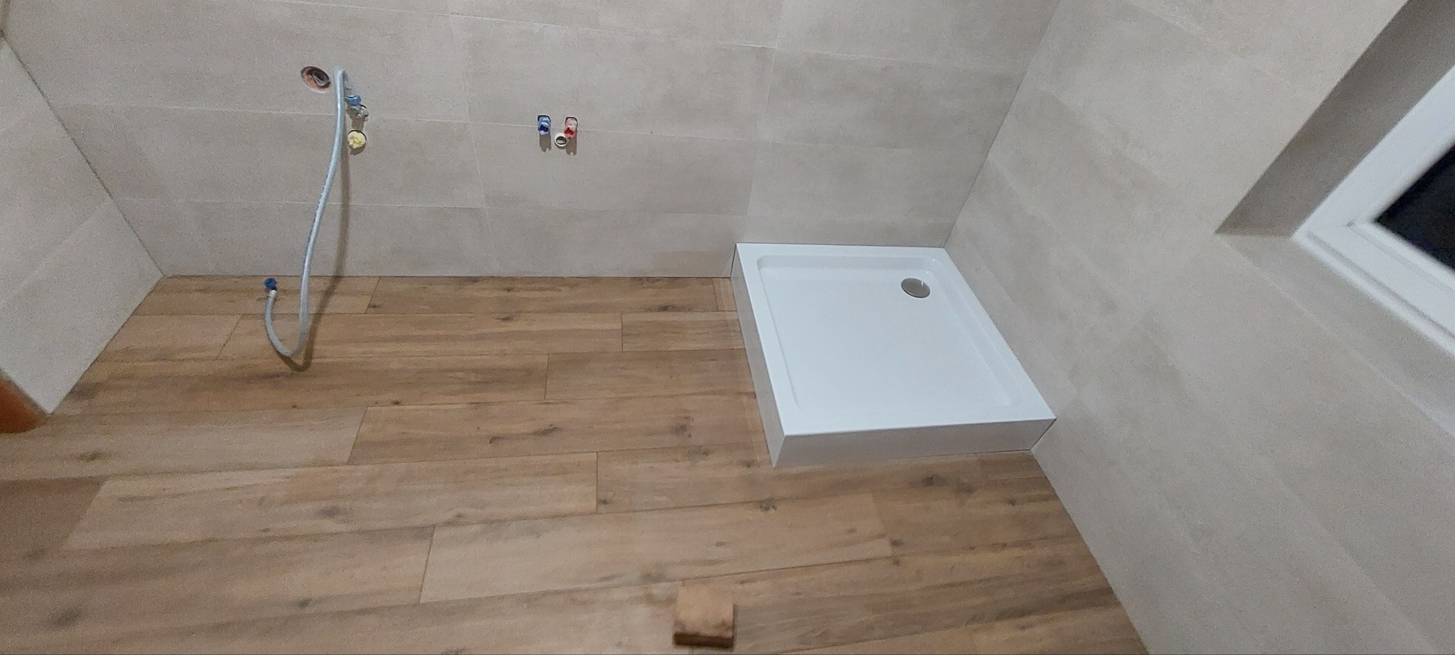 Biały brodzik w rogu wykończonej łazienki z beżowymi płytkami ściennymi i drewnopodobną podłogą. Widoczne przyłącza wodne i wąż prysznicowy.