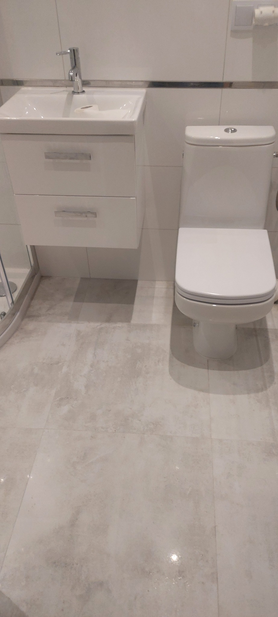 Biała umywalka z szafką i toaleta na tle jasnych, kwadratowych płytek podłogowych i ściennych. Minimalistyczny wystrój łazienki.