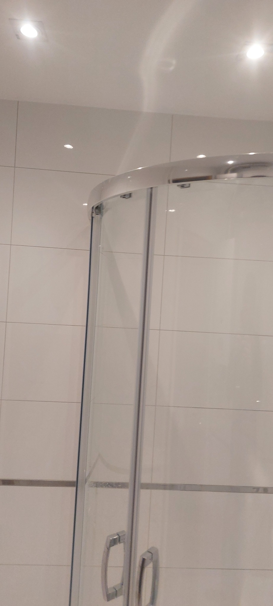 Biała kabina prysznicowa z chromowanymi detalami, widoczna na tle jasnych, kwadratowych płytek. Minimalistyczny design, oświetlenie punktowe w suficie.