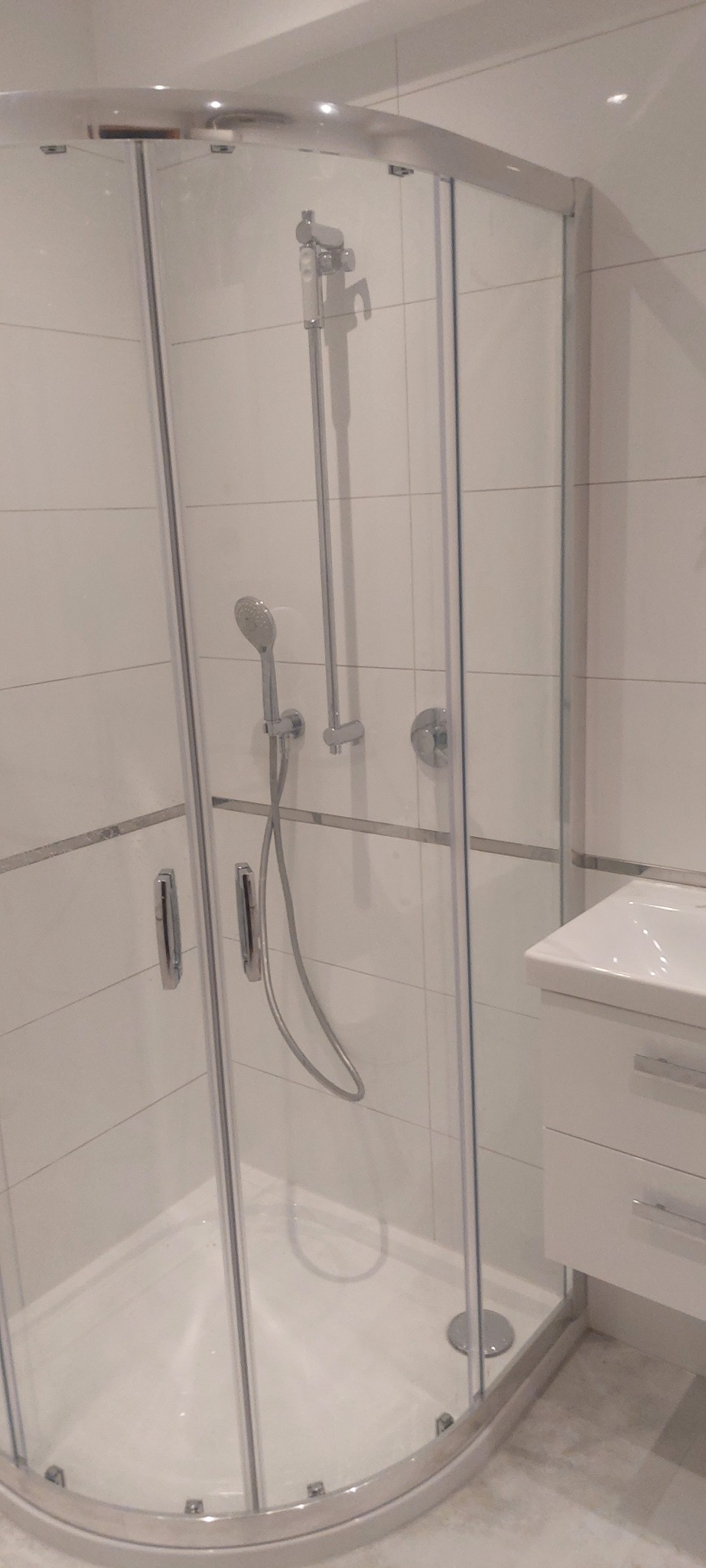 Nowoczesna kabina prysznicowa z chromowanymi detalami i białą armaturą, umieszczona w wykończonej na biało łazience z płytkami i umywalką z szafką.