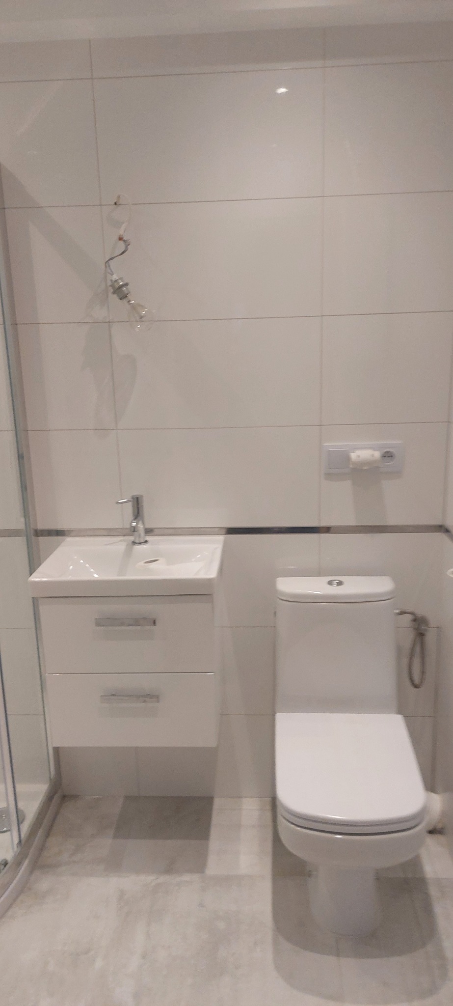 Biała łazienka z umywalką z szafką, toaletą i fragmentem kabiny prysznicowej. Na ścianach białe płytki, widoczna instalacja oświetleniowa.