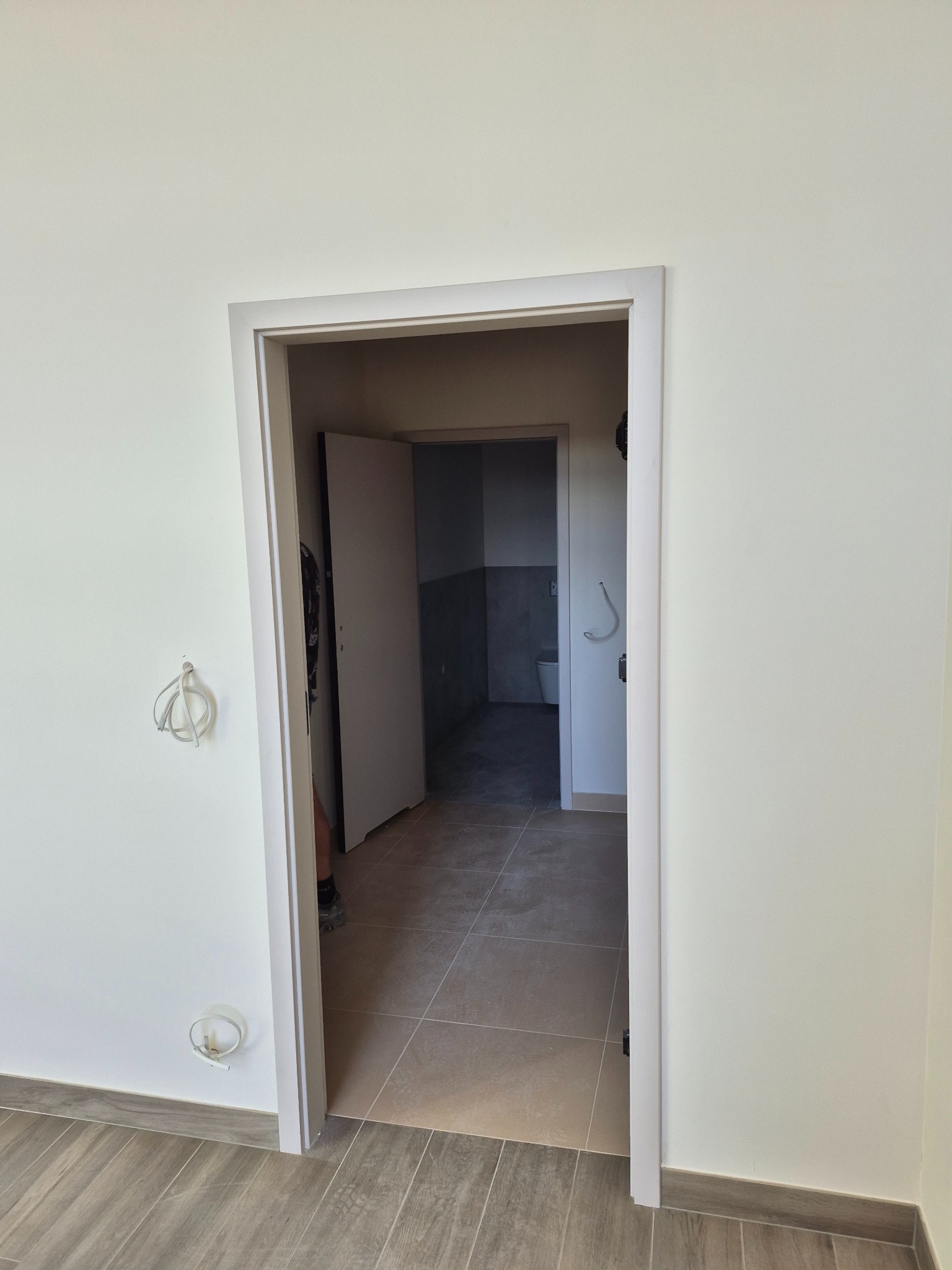 Wykończone ościeże drzwiowe w jasnym wnętrzu, widok na łazienkę z szarą glazurą i białą armaturą. Na ścianie wystają przewody elektryczne.