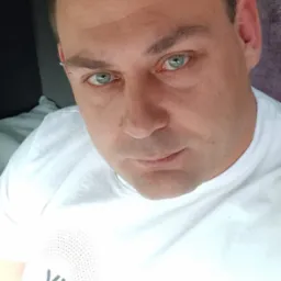 Portret mężczyzny w białej koszulce z napisem 'VISAGEGROUP.', z kolczykiem w brwi, o przenikliwym spojrzeniu zielonych oczu, na rozmytym tle.