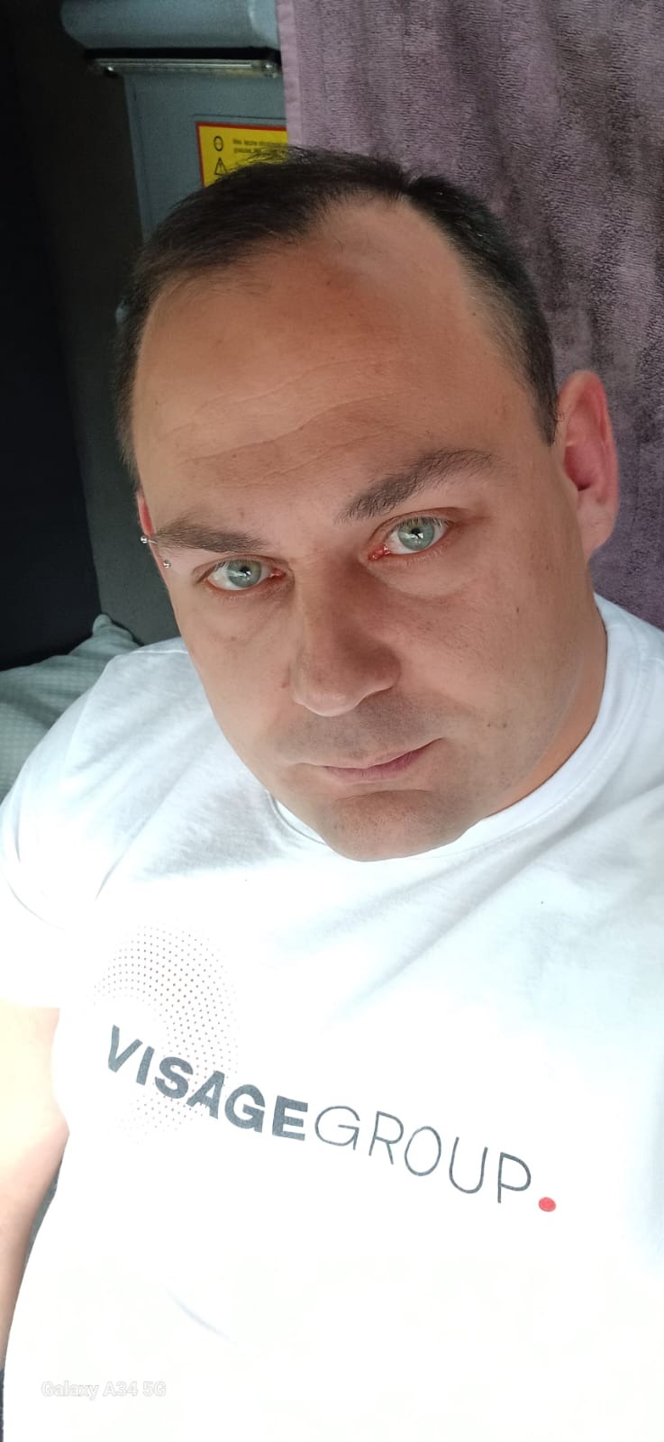 Portret mężczyzny w białej koszulce z napisem 'VISAGEGROUP.', z kolczykiem w brwi, o przenikliwym spojrzeniu zielonych oczu, na rozmytym tle.