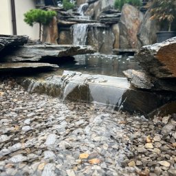 Silva garden - Kaskada wodna w ogrodzie z kamiennymi elementami i roślinnością, z bliska widoczne kamyki i spływająca woda. Elementy domu w tle.