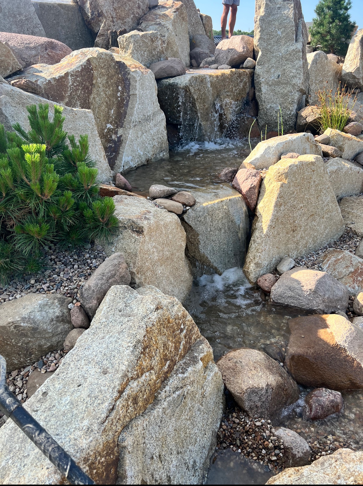 Oczko wodne z kaskadą, otoczone kamieniami i roślinnością. Woda spływa po kamieniach, tworząc mały wodospad. Na górze widać fragment osoby.