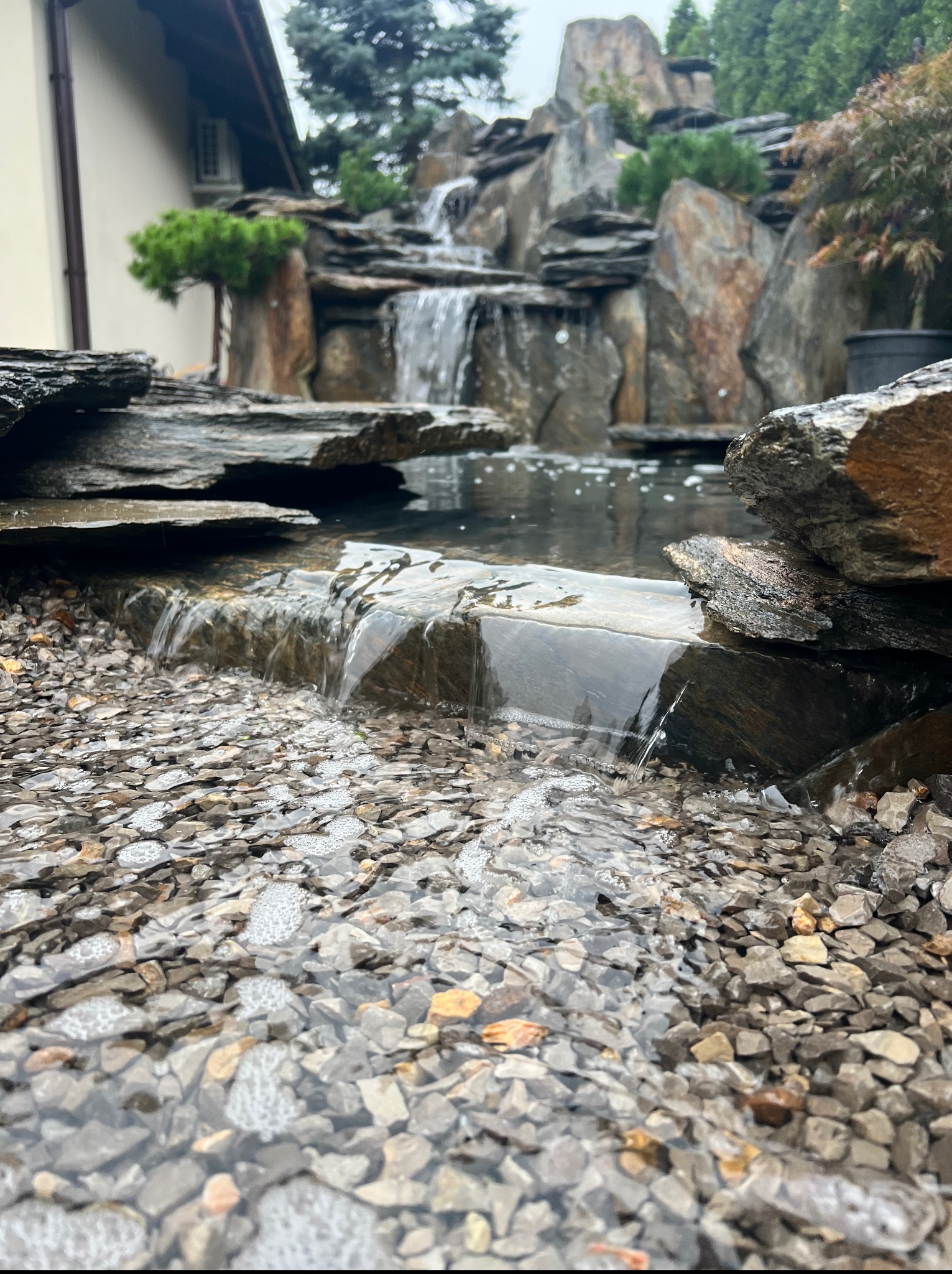 Kaskada wodna w ogrodzie z kamiennymi elementami i roślinnością, z bliska widoczne kamyki i spływająca woda. Elementy domu w tle.