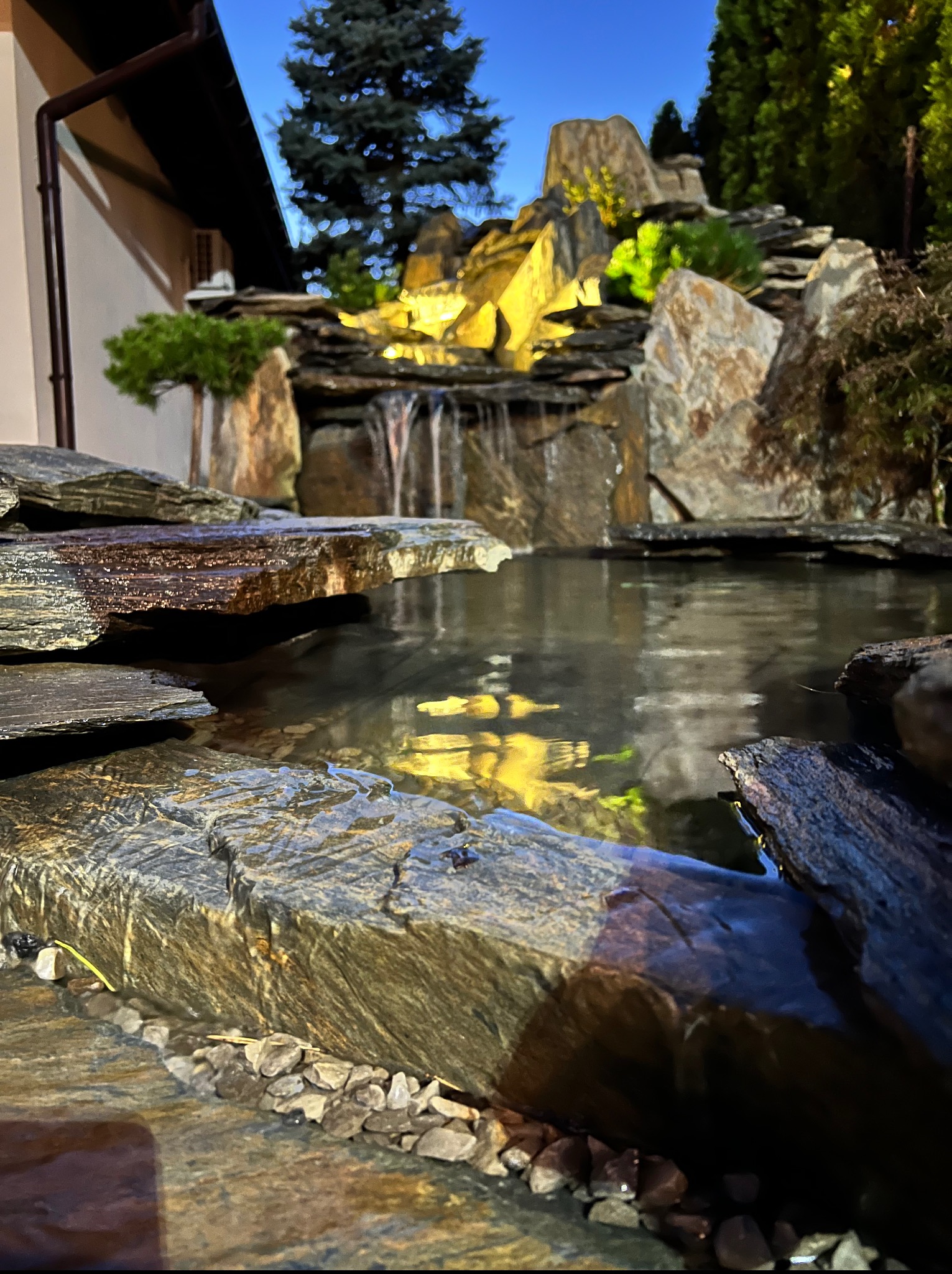 Oczko wodne z kaskadą, podświetlone, otoczone kamieniami i roślinnością. Woda odbija światło, tworząc malowniczy efekt.