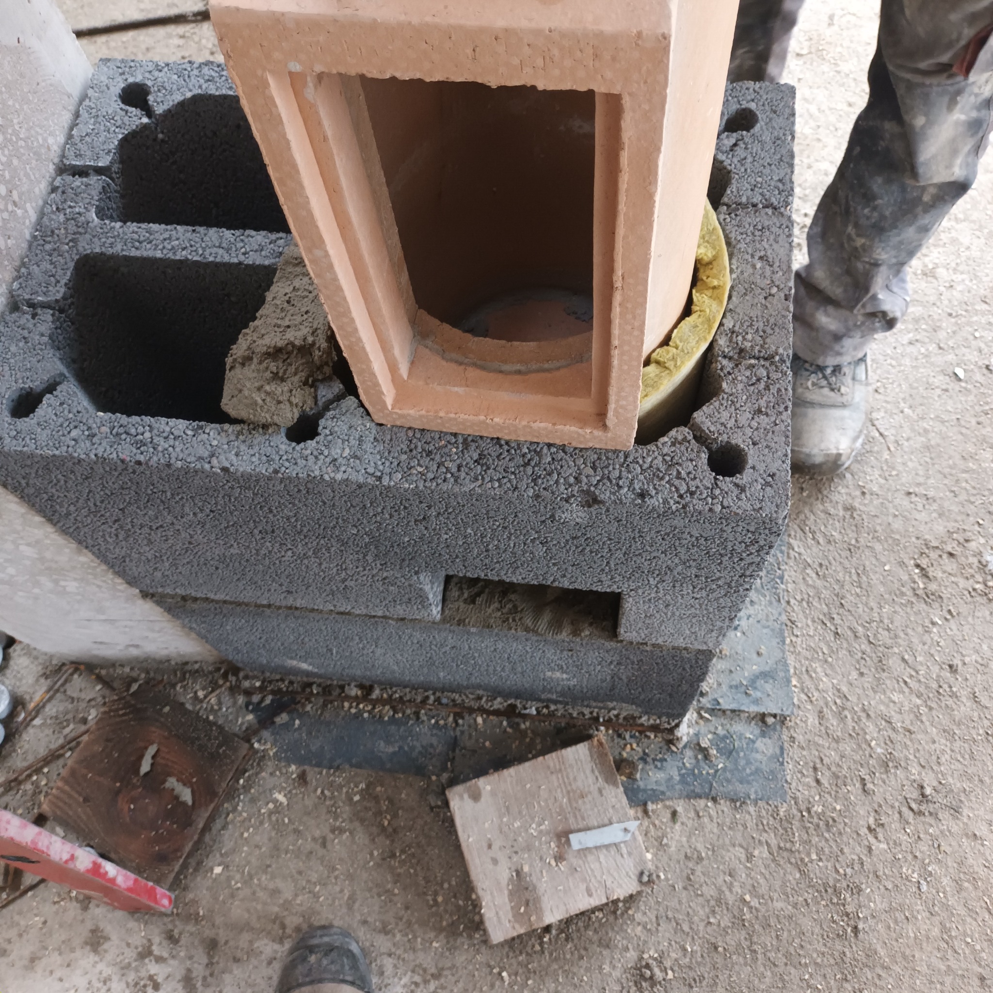 Budowa komina: szare bloczki fundamentowe, ceglana kształtka z izolacją. Widoczne fragmenty robocze i noga pracownika w ubrudzonych spodniach.