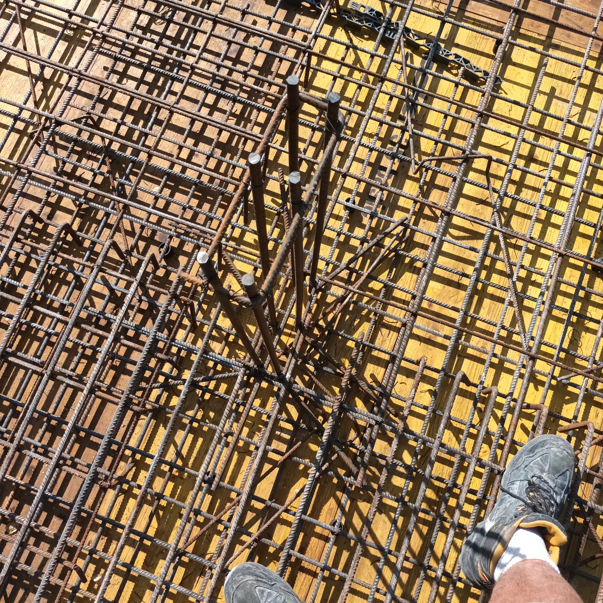 Zbrojenie fundamentów na żółtym szalunku, widok z góry. Stalowe pręty tworzą siatkę, pionowe wzmocnienia. Fragment buta w rogu kadru.