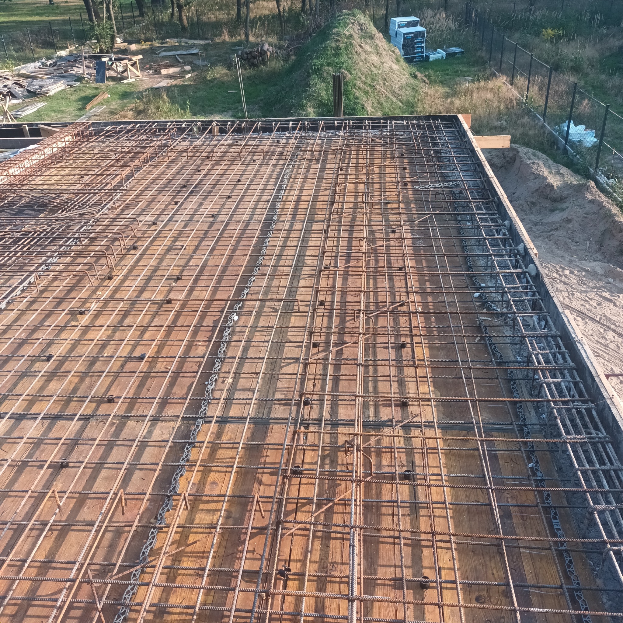 Zbrojenie płyty fundamentowej na deskowaniu, widok z góry. Stalowe pręty tworzą regularną siatkę. W tle teren budowy i elementy konstrukcyjne.