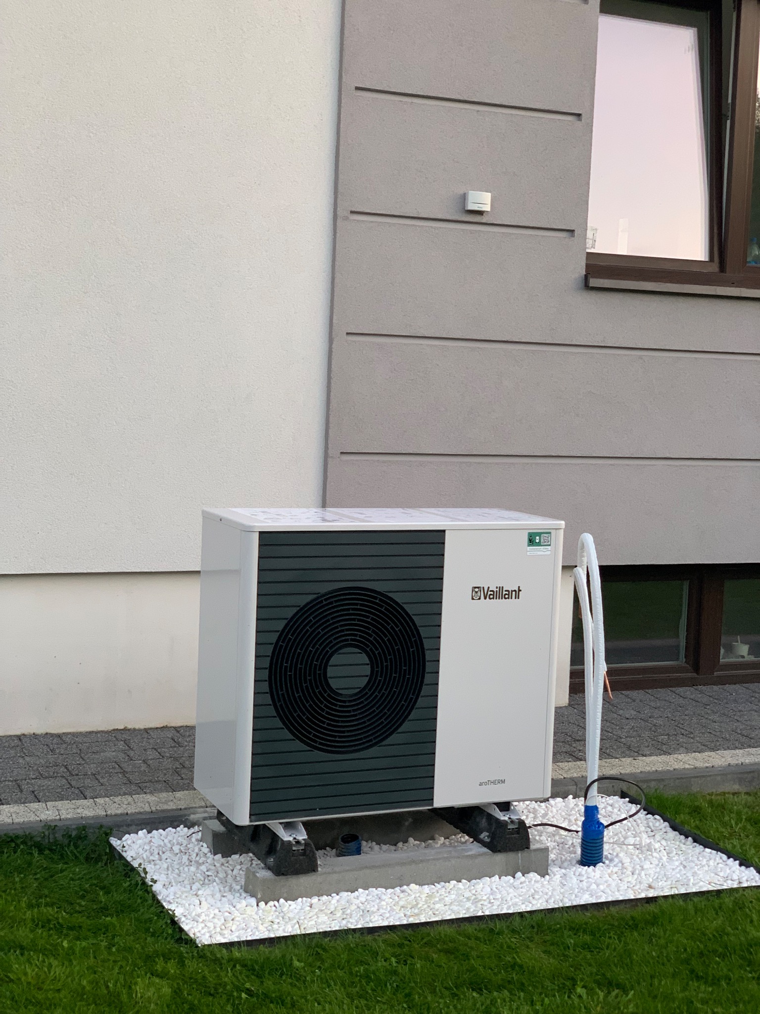 Biała pompa ciepła Vaillant aroTHERM na betonowym fundamencie z białymi kamykami, na tle trawnika i elewacji budynku. Widoczne okno i elementy instalacji.