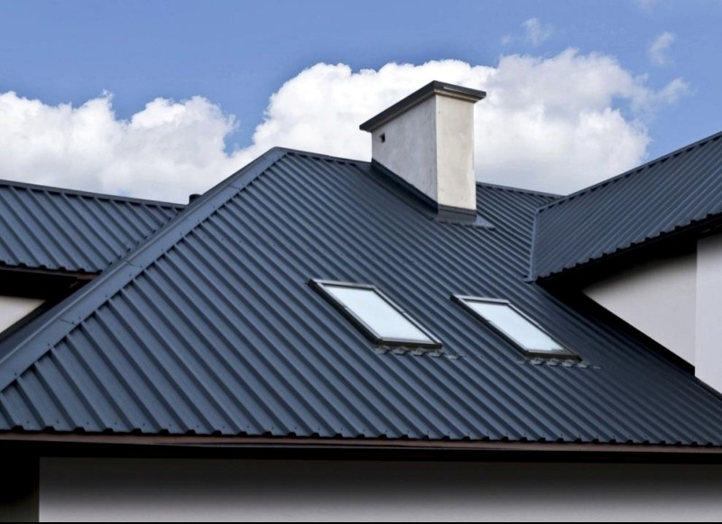 Ciemnoszary dach z blachy trapezowej z dwoma oknami dachowymi i kominem na tle błękitnego nieba z chmurami. Widok z dołu.