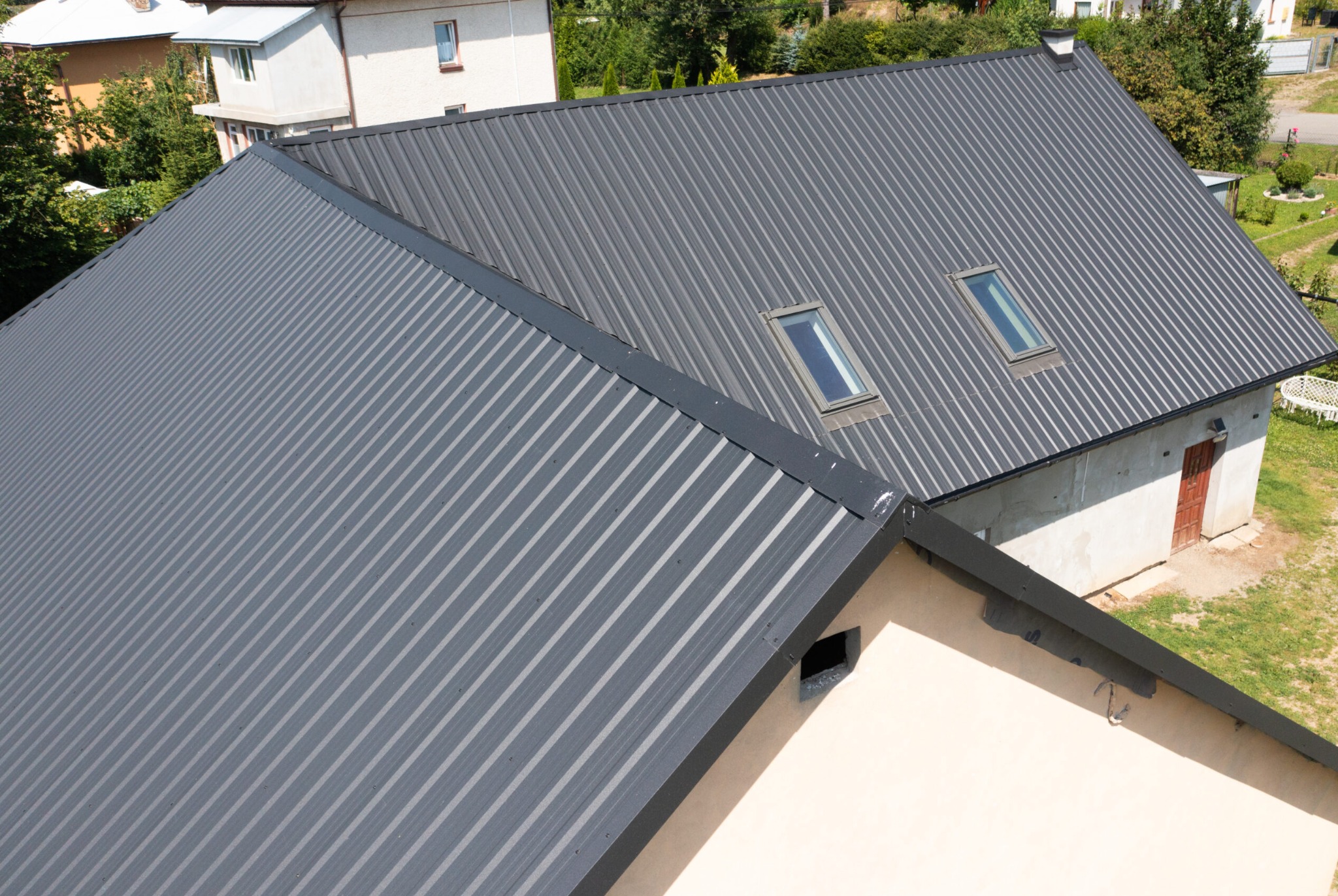Szary dach z blachy trapezowej z dwoma oknami dachowymi, widok z góry, z fragmentami innych budynków w tle.