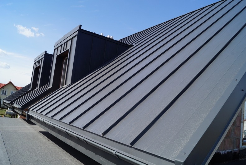 Szary, metalowy dach z oknami dachowymi, widoczny pod kątem, na tle błękitnego nieba. Nowoczesne pokrycie, precyzyjne wykonanie, widoczne obróbki blacharskie.